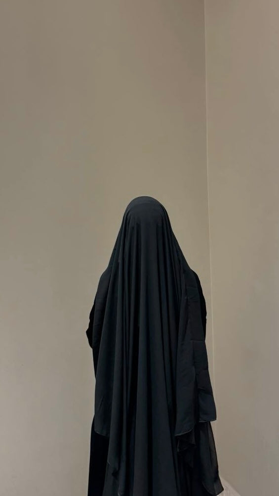 Person wearing a black burqa standing against a plain wall.
