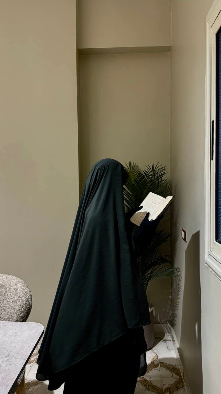 Person wearing a black burqa in a room with beige walls and a plant.