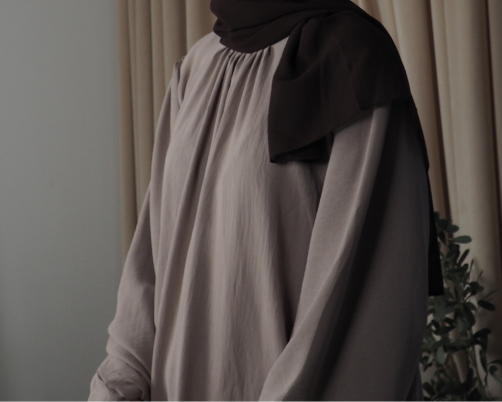 A person wearing a lilac grey abaya with balloon sleeves and pleated details, paired with a black headscarf.
